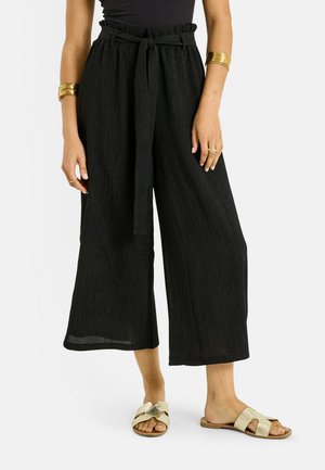 Femme portant un pantalon noir taille haute à jambes larges avec une ceinture en tissu, des bracelets dorés, des bagues et des sandales slides dorées.