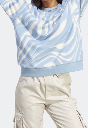 Femme portant un sweat-shirt à motif ondulé bleu et blanc avec un pantalon cargo beige, accessoirisée avec des boucles d'oreilles créoles et un collier chaîne.