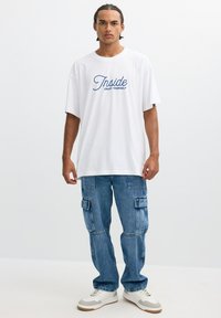 Joven con trenzas que lleva una camiseta blanca que dice "Disfruta de ti mismo", vaqueros cargo azules y zapatillas blancas, de pie frente a un fondo liso.