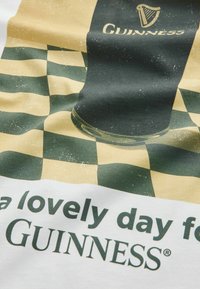 Czarny pint Guinnessa na zielono-beżowym, kratkowanym tle z częściowym napisem "piękny dzień na Guinnessa".