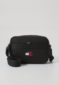 Black fabric crossbody bag with zip closure, featuring a blue, white, and red embroidered logo patch and a detachable strap.