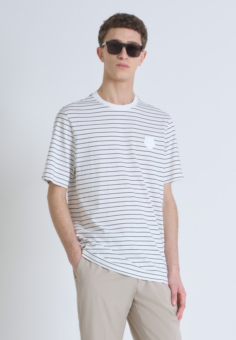 Camiseta de rayas blancas con mangas cortas, que presenta rayas horizontales negras y un pequeño emblema blanco en el pecho. Combinada con pantalones beiges.