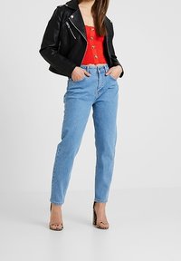 Chaqueta de cuero sintético negra, top de punto rojo con botones, vaqueros de tiro alto azules, y como calzado, tacones transparentes con un patrón texturizado.