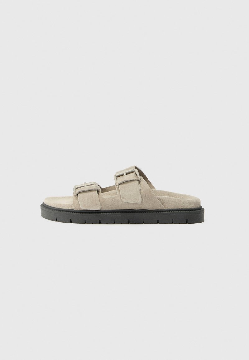 Sandale din suede de culoare gri deschis, cu două curele ajustabile și catarame argintii. Prezintă o talpă groasă din cauciuc negru cu textură.