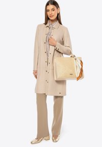 Beige leren jas met drukknoopjes, gestreept shirt eronder, beige broek, gouden schoenen en een textuur crème handtas met een bruine accentlint.