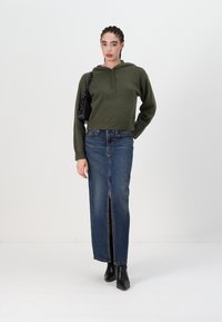 Sötétzöld, cropped kapucnis pulóver, klasszikus farmer elülső részen hasítással, és fekete bokacsizma, ami egy laza, mégis modern sziluettet alkot.