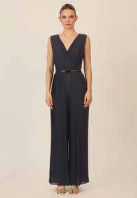 Mono de marina con escote en V profundo y diseño sin mangas, con tela plisada y un cinturón negro ajustado en la cintura. Tacones dorados completan el look.