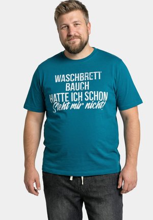 Bärtiger Mann lächelt, trägt ein tealfarbenes Hemd mit deutschem Text "Waschbrett Bauch hatte ich schon steht mir nicht!" und dunkle Jeans, steht vor weißem Hintergrund.