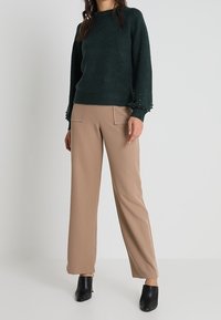 Tumma vihreä neulepusero, jossa on pyöreä pääntie ja pitkät hihat, sisältää helmikoristeet hihoissa. Yhdistetty vaaleanbeigeihin leveälahkeisiin housuihin ja mustiin avokkaisiin.