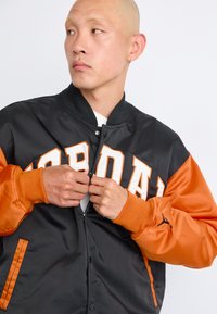 Man with shaved head zips up black and orange Jordan jacket with large white lettering on front, against plain white background.