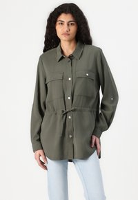 Olijfgroene shirt met twee borstzakken, knopen voorkant en verstelbare tailleband. Lange mouwen met knopen manchetten, gemaakt van een gladde stof.