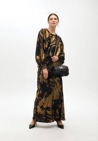 Lange, vloeiende jurk in een gouden en zwarte tie-dye patroon; heeft uitlopende mouwen en een ronde halslijn. Accessoire: zwarte bonten handtas met kettingband.