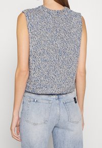Bezrekawowy dzianinowy top w kolorze niebieskim i beżowym z teksturowanym wzorem i wysokim dekoltem. Połączony z jasnoniebieskimi dżinsami z tylnymi kieszeniami.