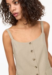 Débardeur beige à côtes avec décolleté en cœur, quatre boutons sur le devant et fines bretelles. Le tissu a une texture douce et un léger élancement.