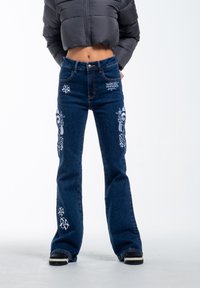 Flared denim jeans in dark blue with intricate white embroidery of flowers and skulls on the legs; medium weight fabric, visible stitching.