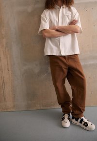 Kortärmad vit skjorta med knapp och ficka, matchad med bruna, löst sittande byxor och vita sneakers med blå accenter.