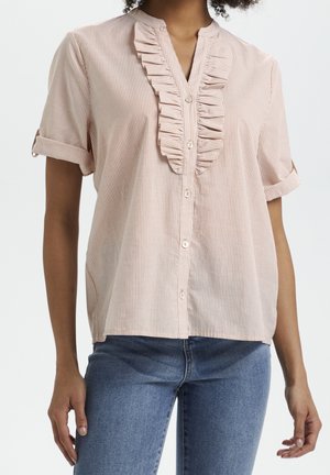 Vrouw draagt een lichtroze blouse met korte mouwen, knoopsluiting, verticale strepen en ruches, gecombineerd met blauwe spijkerbroek.