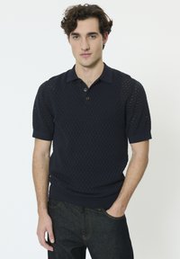 Polo manches courtes bleu marine présentant un design en maille ouverte texturée, patte à deux boutons et poignets côtelés. Porté avec un jean foncé.