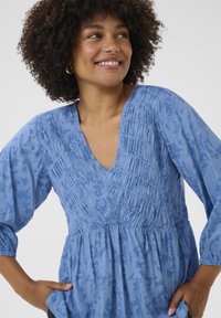 Kék textil blúz V-nyakkal, ráncolt részletekkel és háromnegyedes ujjakkal. Az anyag finom virágmintával és puha textúrával rendelkezik.