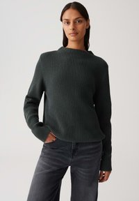 Donna con lunghe capelli scuri, che indossa un maglione a coste scuro e jeans neri sbiaditi, in piedi con una mano in tasca davanti a uno sfondo neutro.