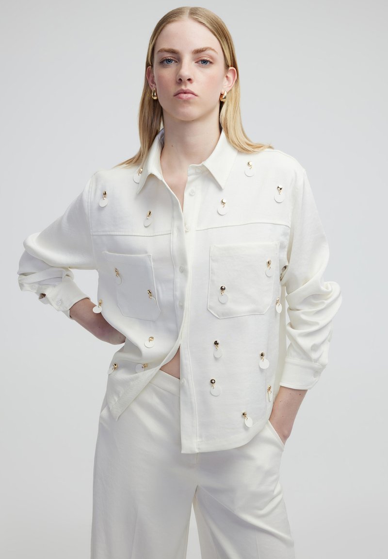 Witte blouse met gouden accenten en decoratieve ronde hardware, voorzien van twee voorzakken en een ontspannen pasvorm, gestyled met opgerolde mouwen.