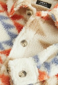 Veste en polaire douce avec un motif géométrique multicolore, boutons beige et col, avec une étiquette indiquant "NEXT Made in Bangladesh."