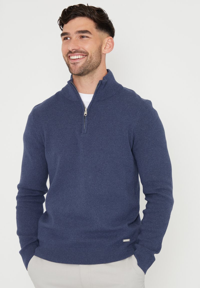 Marineblauer Strickpullover mit Reißverschluss, hohem Kragen, gerippten Bündchen und Saum. Weiche Textur mit kleinem Logoakzent nahe dem unteren Rand.