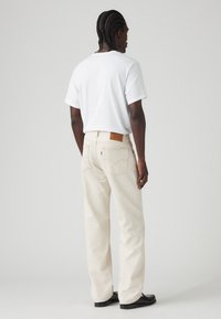 Tricou alb asortat cu blugi bej deschis. Blugii au o etichetă din piele distinctivă pe talia din spate și buzunare clasice la spate. Pantofi negri.