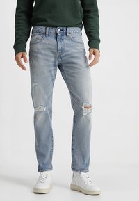 Jeans de mezclilla azul claro con detalles rasgados, que presentan un corte recto, cinco bolsillos y un cierre de botón. Combinados con zapatillas blancas.