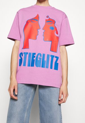Lila katoenen T-shirt met een grafisch ontwerp van twee abstracte gezichten in rood en blauw, met het woord "STIEGLITZ" in vette blauwe letters eronder gedrukt.