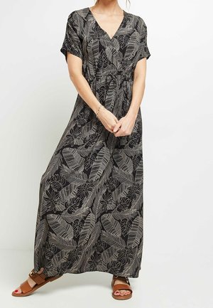 Kobieta ubrana w czarną i beżową sukienkę maxi w liściasty wzór, z krótkimi rękawami i brązowe płaskie sandały, stojąca na tle jednolitego białego tła.