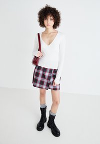 Weißes Langarmoberteil mit V-Ausschnitt, kombiniert mit einem karierte Minirock in Rot und Schwarz. Schwarze Schnürstiefel, graue Socken und eine rote Handtasche.