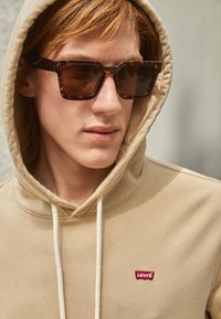 Beige hettegenser med snor og rødt Levi's-logo. Brune skilpaddemønstrede solbriller med firkantet ramme. Jevn tekstur, nærbilde.