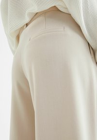Beige byxor med en slät yta, utrustade med en bakficka. Parat med en ribbad vit tröja. Tyget är lätt och mjukt.
