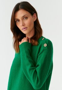 Pull vert avec une finition texturée, des poignets côtelés et trois boutons décoratifs sur l'épaule. Tissu doux et coupe décontractée.