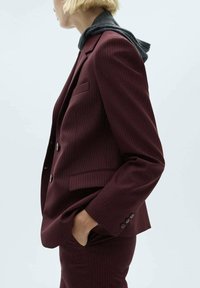 Personne portant un costume rayé rouge foncé avec les mains dans les poches et un sweat à capuche sombre en dessous, faisant face à gauche sur un fond uni.