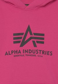 Felpa rosa con un grafico nero raffigurante una "A" circondata da ali e la scritta "ALPHA INDUSTRIES, KNOXVILLE, TENNESSEE, U.S.A."