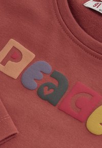 Close-up van een rode sweatshirt met kleurrijke reliëfletters die "peace" spellen, met een hartvorm binnen de letter "a."