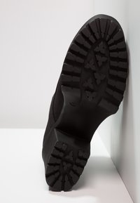 Semelle de botte noire avec un motif de crampons profond, positionnée verticalement contre un mur et une surface blancs, affichant la taille 37 sur la semelle.
