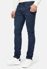 Vaqueros de mezclilla azul oscuro con corte ajustado, diseño de cinco bolsillos y detalles de costura visibles. Combinados con zapatillas blancas.