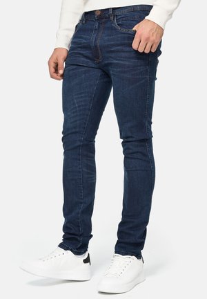 Dunkelblaue Jeans aus Denim mit Slim-Fit, Fünf-Taschen-Design und sichtbaren Nahtdetails. Kombiniert mit weißen Turnschuhen.