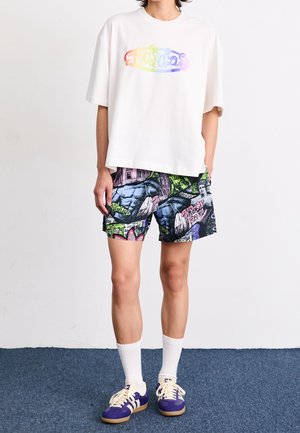 Person iført en hvid oversized T-shirt med regnbuefarvet tekst og farverige grafiske tryk shorts, hvide sokker og lilla sneakers.
