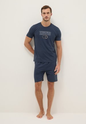 Uomo in piedi a piedi nudi contro un muro bianco che indossa pantaloncini e t-shirt blu navy con la scritta "WEEKEND MODE ON" e un'icona di interruttore.