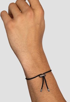 Schwarzes geflochtenes Armband mit metallischen Akzenten, ausgestattet mit einem schiebbaren Verschluss. Getragen an einer Hand mit sichtbarem Hautbild und Haar.