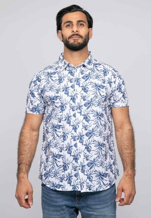 Homme aux cheveux courts et foncés avec une barbe, portant une chemise blanche à manches courtes avec un imprimé floral bleu et un jean bleu, debout devant un fond gris.