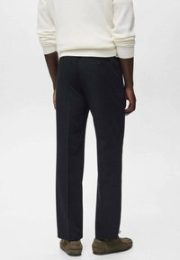 Homme de dos portant un pantalon noir sur mesure associé à un pull blanc et des chaussures en daim marron sur fond uni.