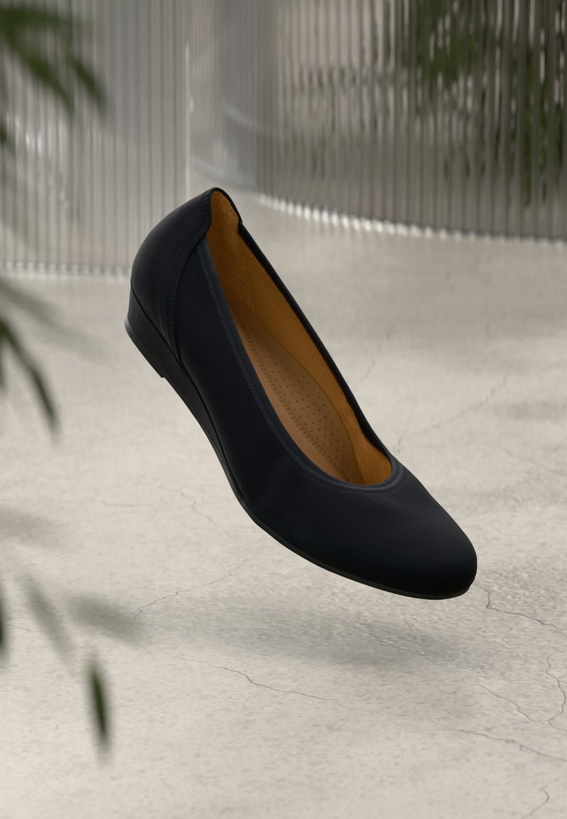 Ballerine en cuir noir avec semelle intérieure rembourrée beige, présentée en suspension au-dessus d'un sol en béton texturé, avec des plantes et du verre flous en arrière-plan.