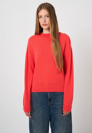 Jonge vrouw met lang, stijl haar, gekleed in een rode trui en blauwe spijkerbroek, staand tegen een effen lichte achtergrond.