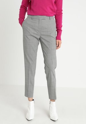 Femme portant un pantalon gris sur mesure jusqu'aux chevilles, un haut rose à manches longues et des bottines blanches, debout les mains dans les poches.