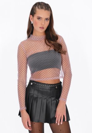 Ung kvinde med langt hår iført en gennemsigtig lyserød polkaprikbluse over en grå crop top, sort plisseret lædernederdel og matchende polkaprikstrømpebukser.